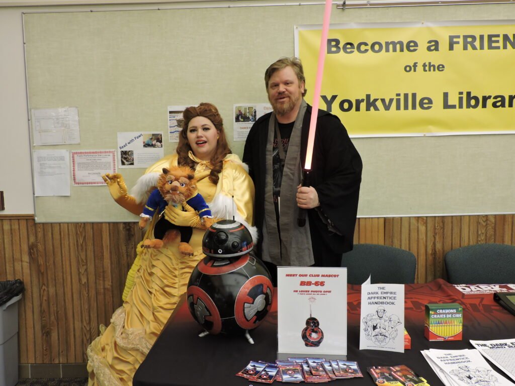 A women dressed as Belle from Beauty and the Beast holds a Beast doll stands next to a man with a lightsabre