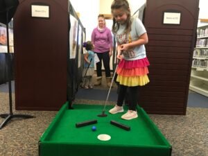 Young girl putts into mini golf hole while younger girl and mom wait their turn.