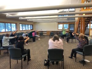 Eleven women sit in chairs in a semi circle stretching their arms above their head.
